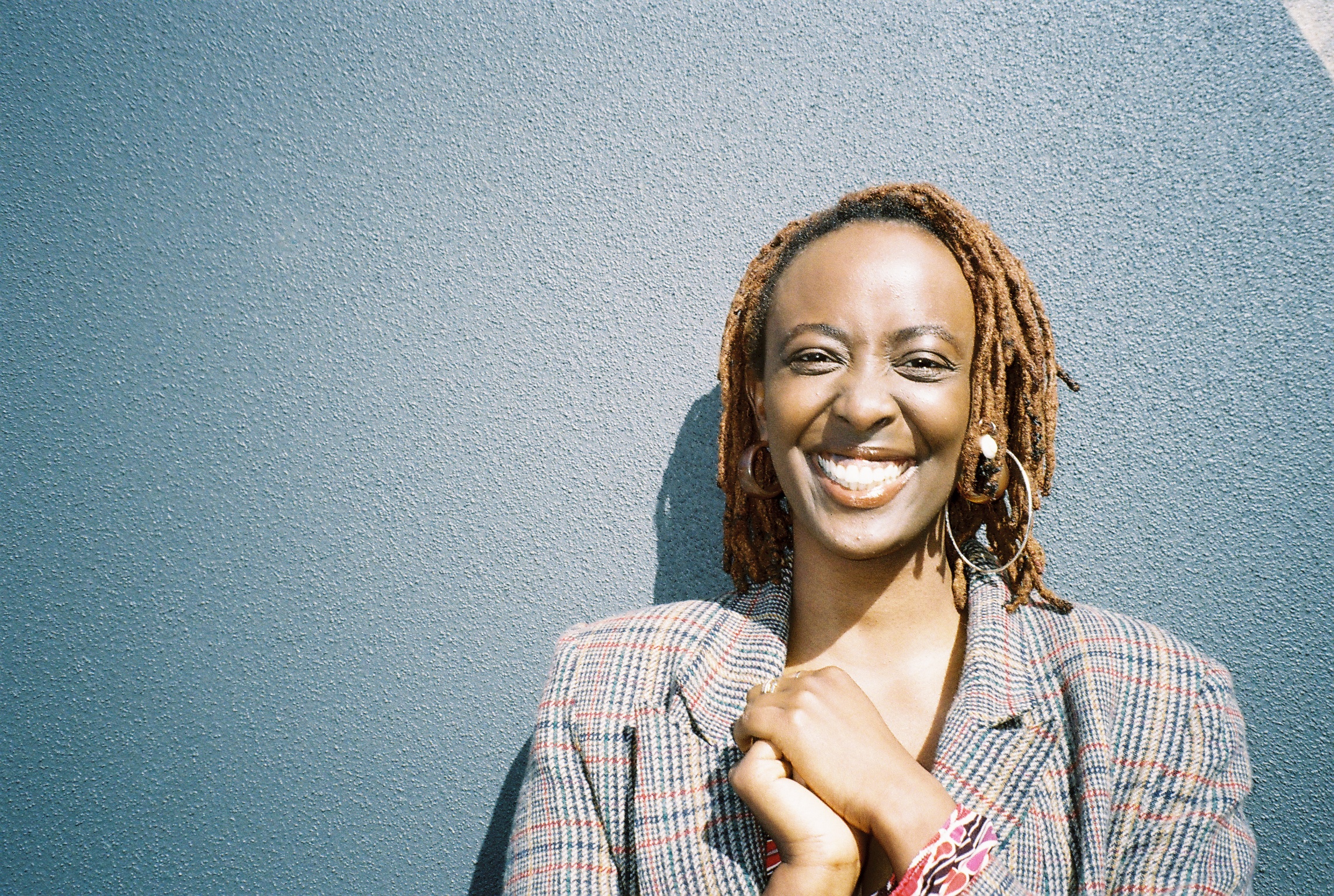 A portrait of a Rwandan woman in her mid-twenties with shoulder length brown dreadlocks smiling at the camera. She is wearing brown and silver earrings and a chequered jacket with blue, red, green, brown lines