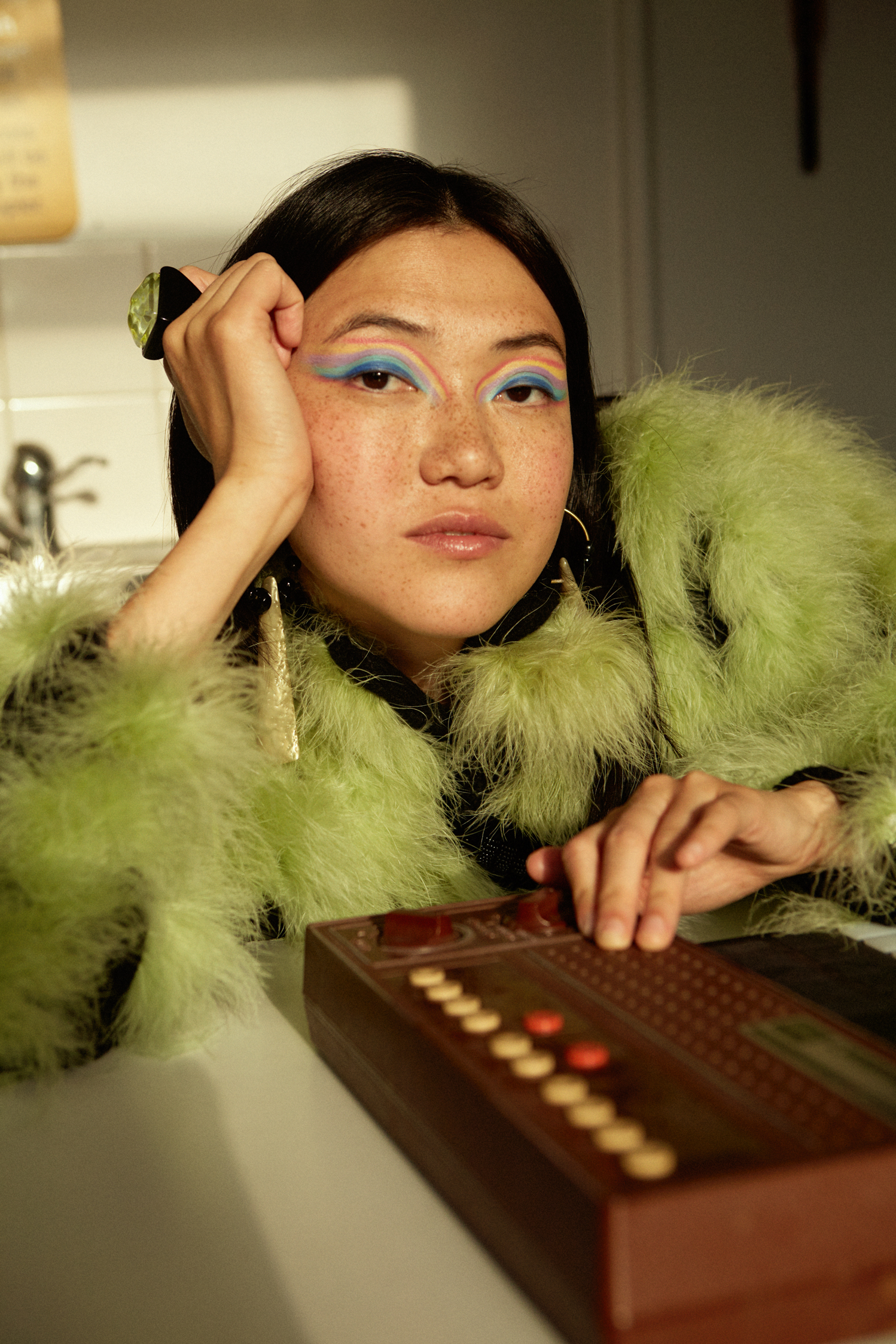 Woman with short dark hair and colourful eyeshadow in a fluffy green jumper casually leans on a table in a kitchen playing with an instrument.