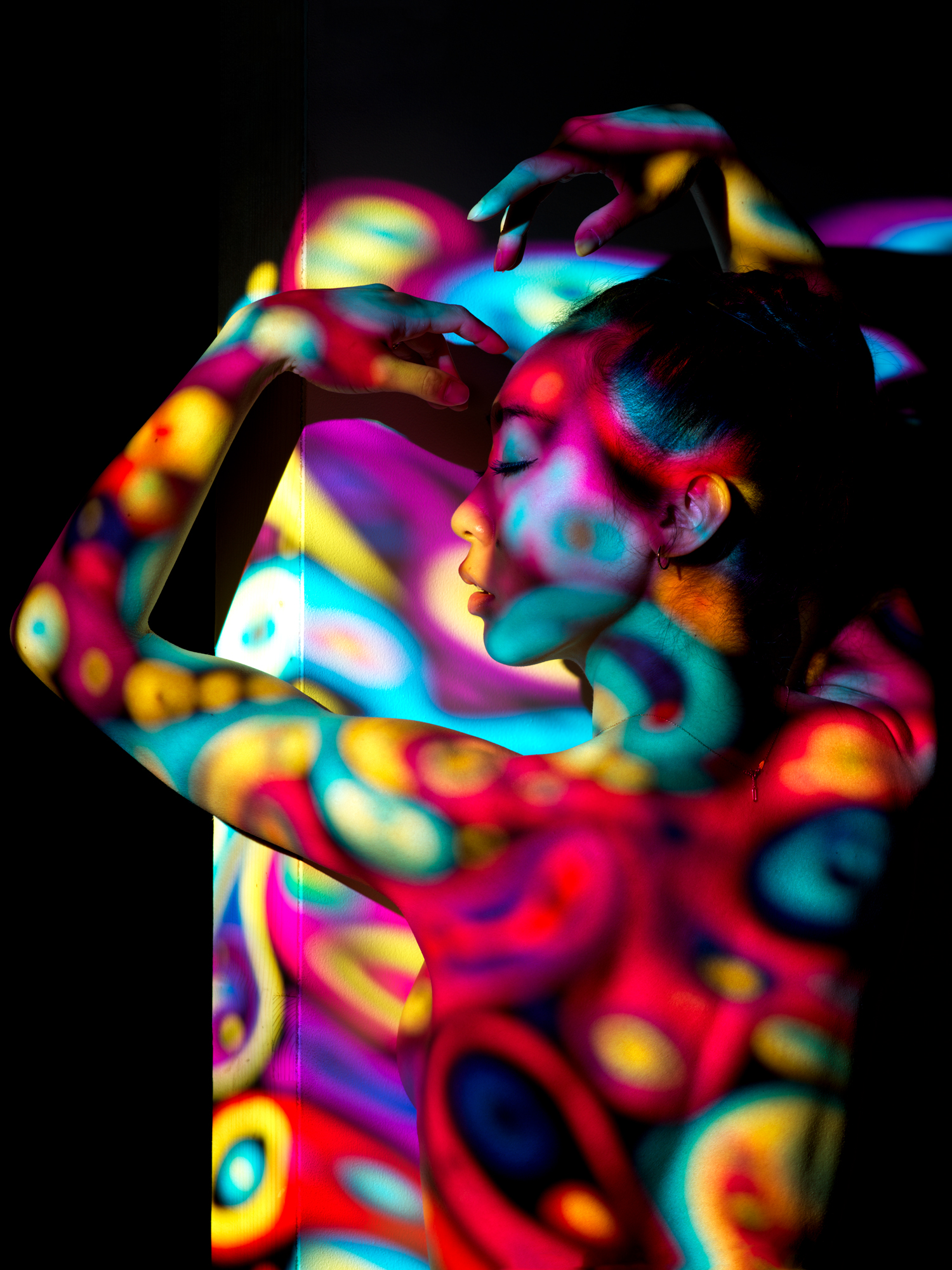 An image of a woman with black hair and arms raised covered by a multi coloured projection all over her body