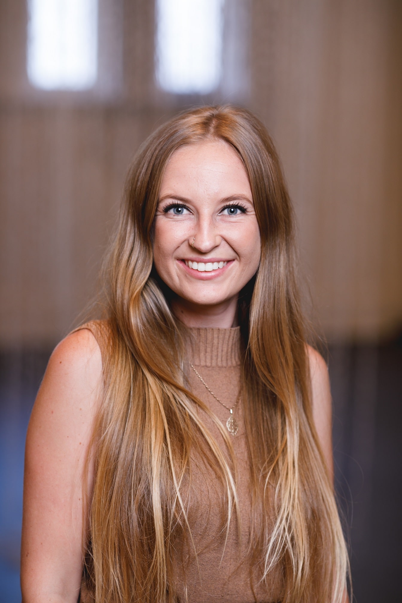 A woman with long blonde hair smiling with a blurred background of a room with an indistguishable hanging artwork behind