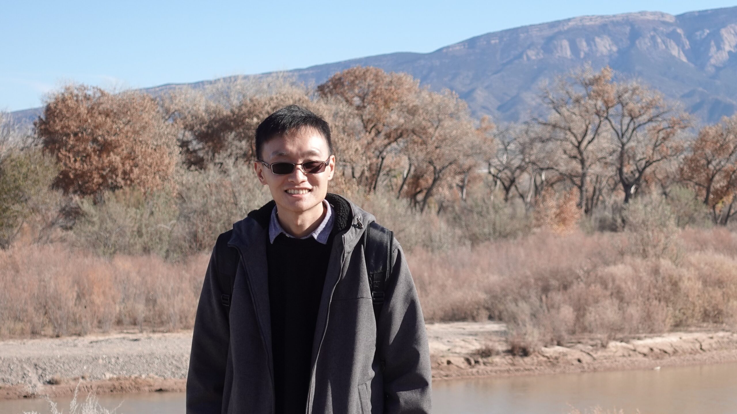 This is the bust of the speaker, his hair is short and black, he's wearing a dark colour coat and a pair of sunglasses. The photo is taken by the river, with autumn forest and mountain in the background.