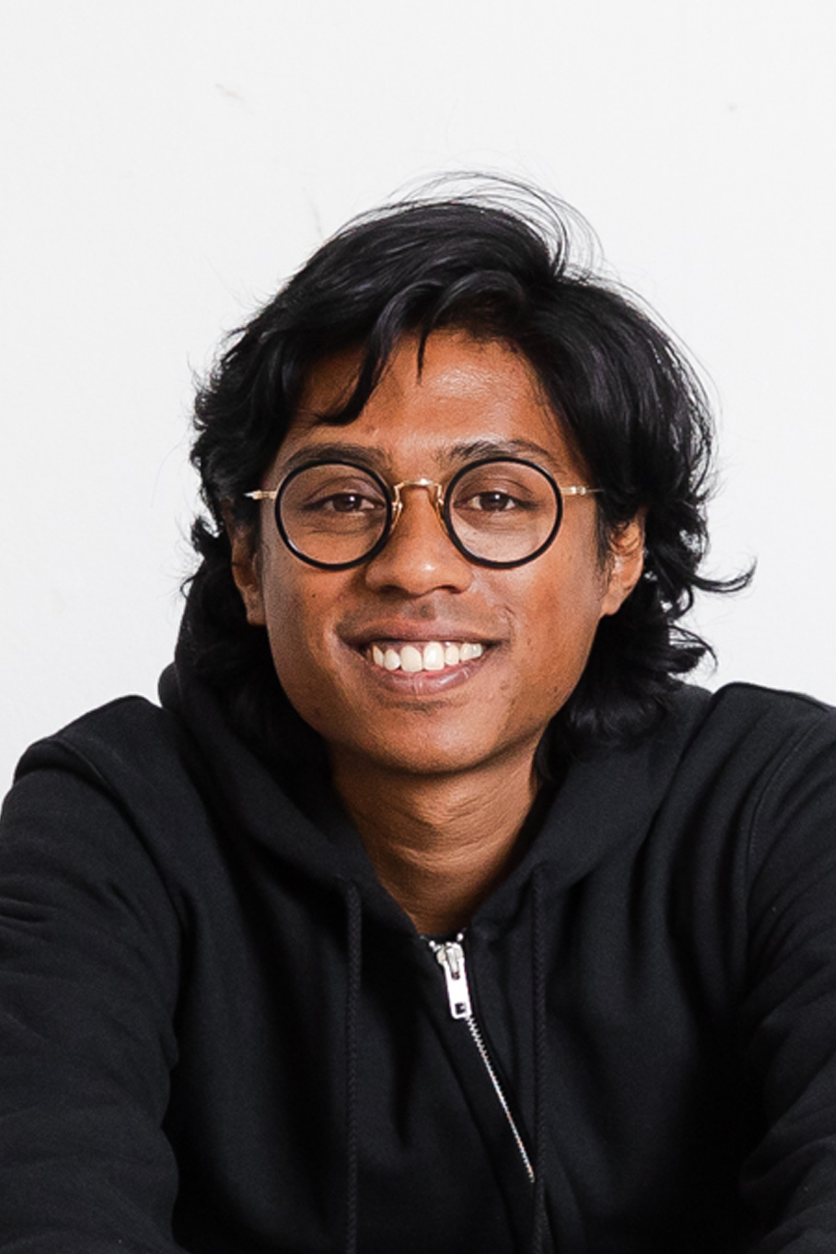 An image of a man in black rimmed glasses, wearing a black hodie with a white background and smile on his face