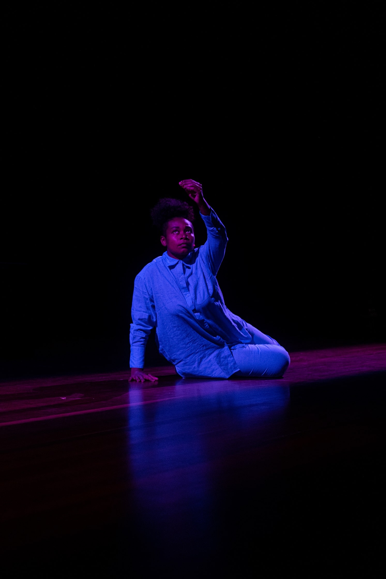 A person sits on a stage floor with a purple toned spotlight on them. They are wearing all blue and one hand is raised.