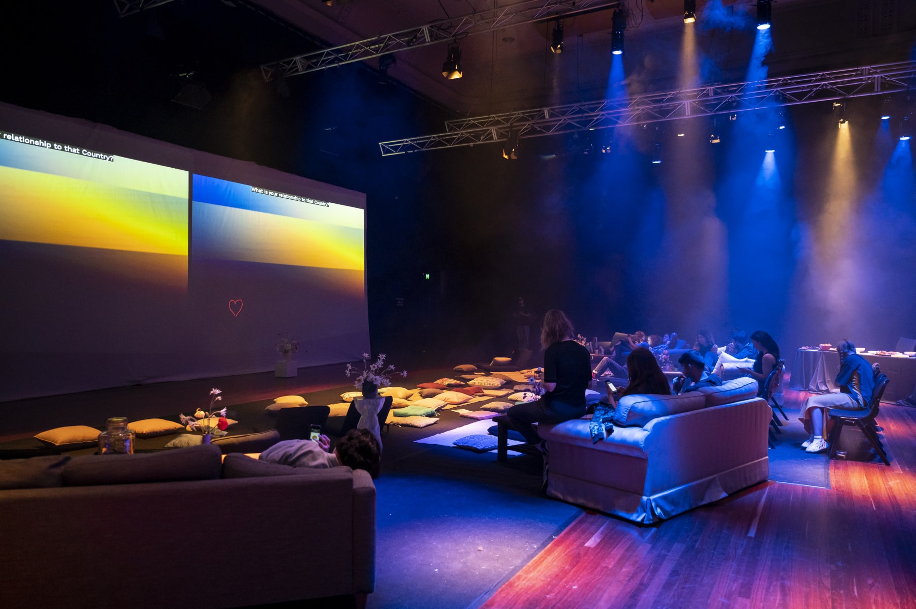 A group of people in a dimly lit room sitting on couches and cushions. They are looking at their phones and responding to prompts on a large screen. There is haze and coloured lights.